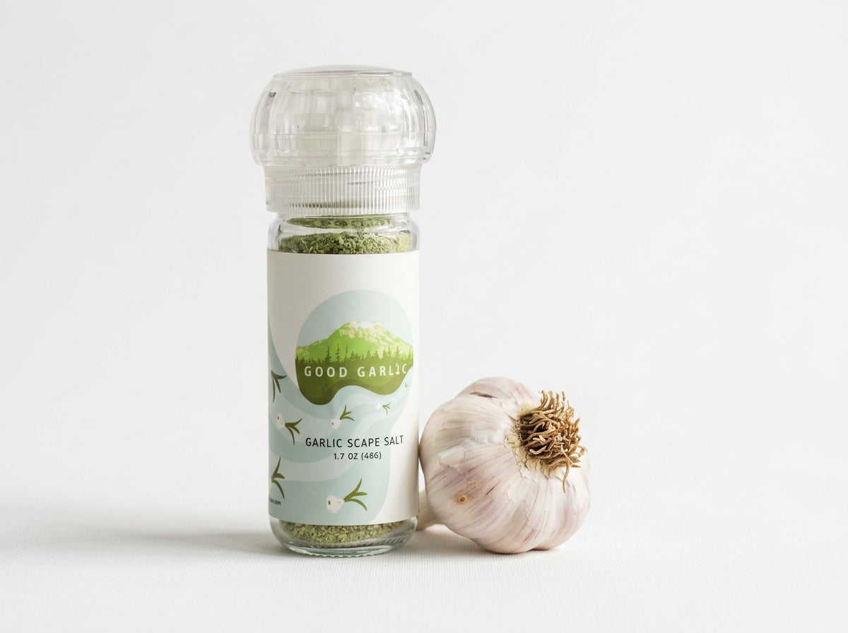 A bottle of Good Garlic Garlic Scape Salt with a clear cap and a white label, shown next to a whole garlic bulb on a clean white background.
