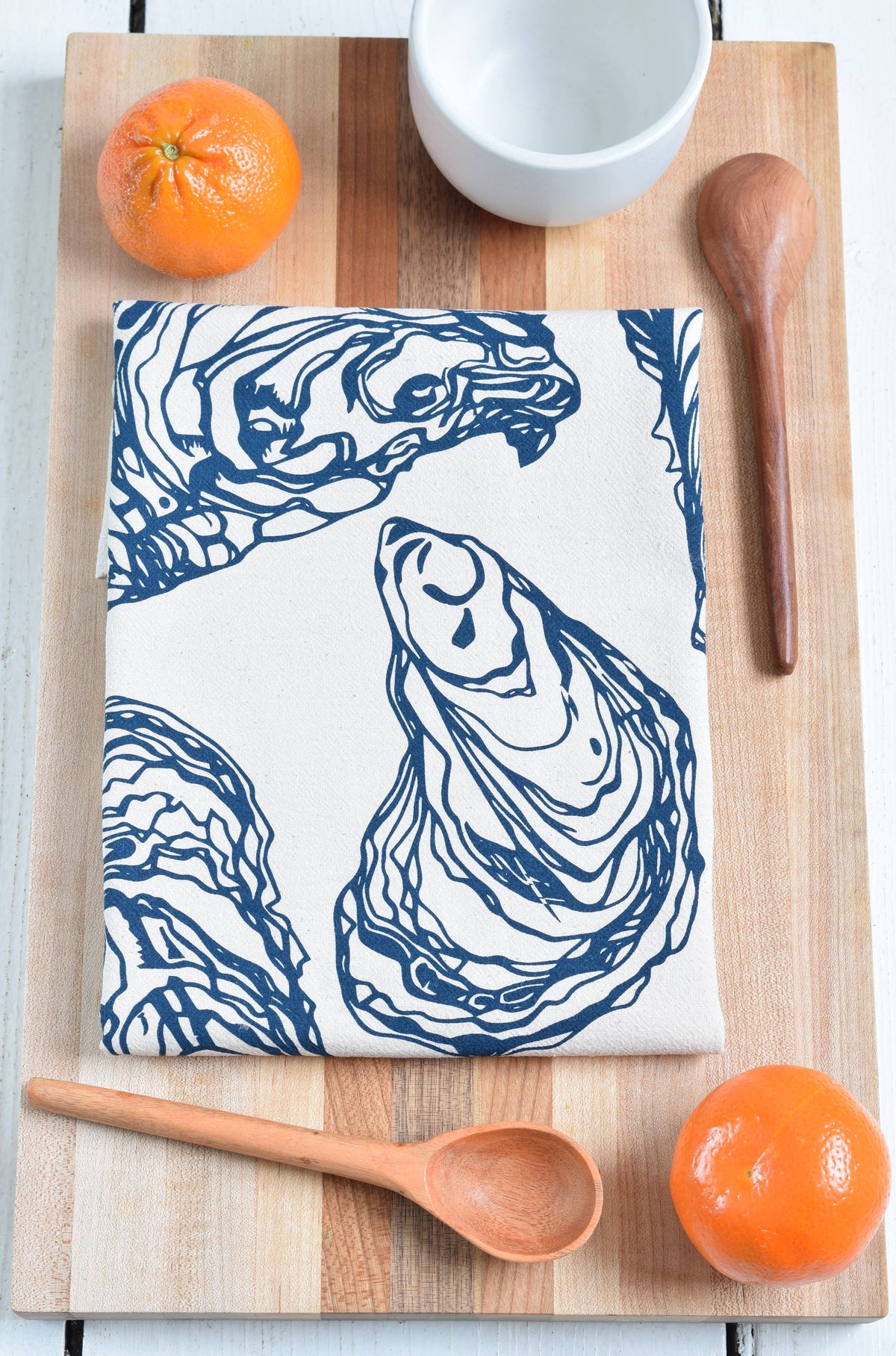 Oyster print towel on a cutting board with oranges wooden spoons and a cup.