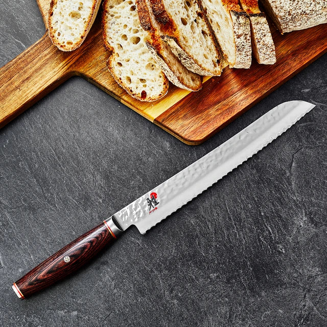 Miyabi Artisan Bread Knife on textured black background with a cutting board with sliced bread.