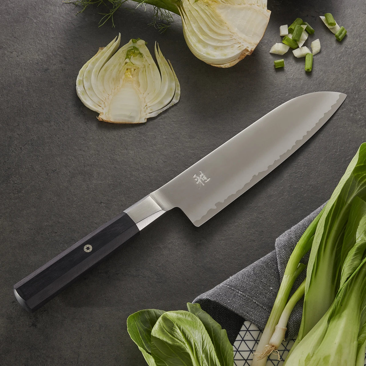 Myabi Koh Santoku on textured black background with various vegetables..