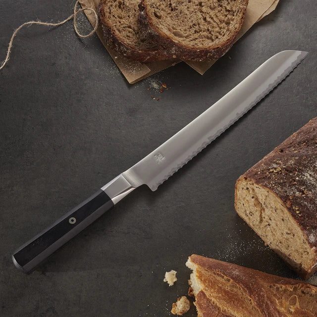 Miyabi Koh Bread knife on textured black background with bread.