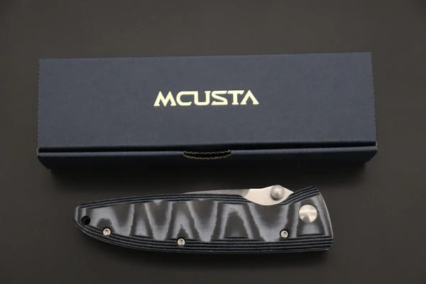 An MCUSTA folding pocket knife with a textured black handle and silver metal hardware, displayed below its dark navy blue presentation box on a dark background.