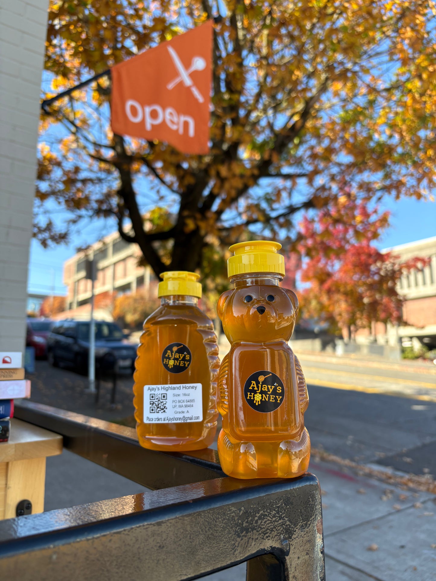 Two bottles of Ajay's Honey, one bear-shaped and one rectangular, filled with golden honey and standing on a railing outdoors with a street and autumn tree in the background.