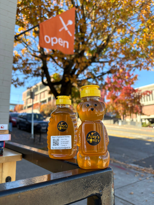Two bottles of Ajay's Honey, one bear-shaped and one rectangular, filled with golden honey and standing on a railing outdoors with a street and autumn tree in the background.