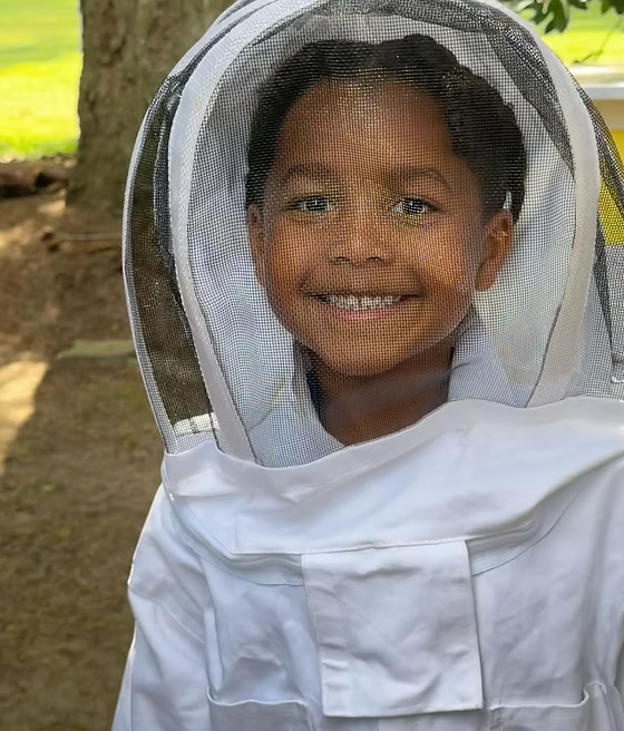 A smiling young boy wearing a white beekeeping suit with a mesh face veil, photographed outdoors in front of a tree.