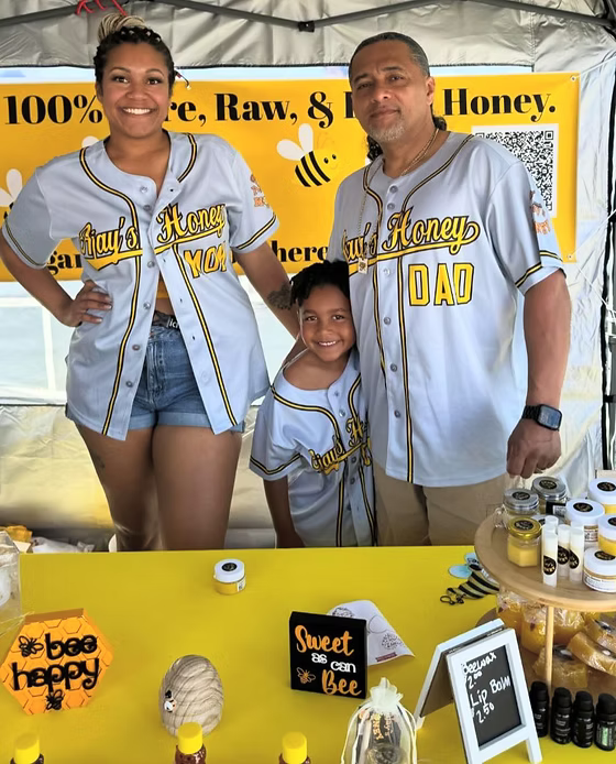A family of three posing together at a honey booth under a tent, wearing matching white baseball-style jerseys with yellow and black lettering that reads 'Sweet Honey'. The table in front displays various honey products, jars, and bee-themed signs.