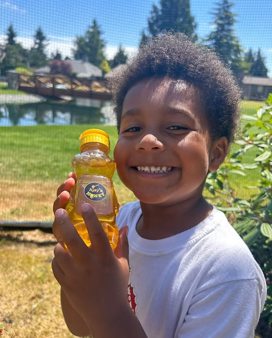 A smiling young boy holding a bottle of Nature's Organic Raw Honey outdoors in front of a pond and greenery.