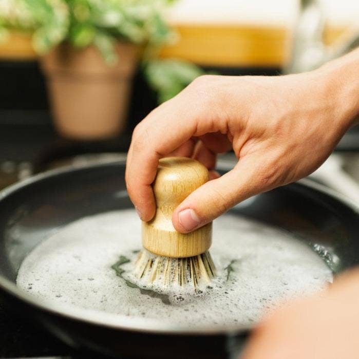 Hand using a wooden brush to scrub a pot with soapy water in a kitchen setting.