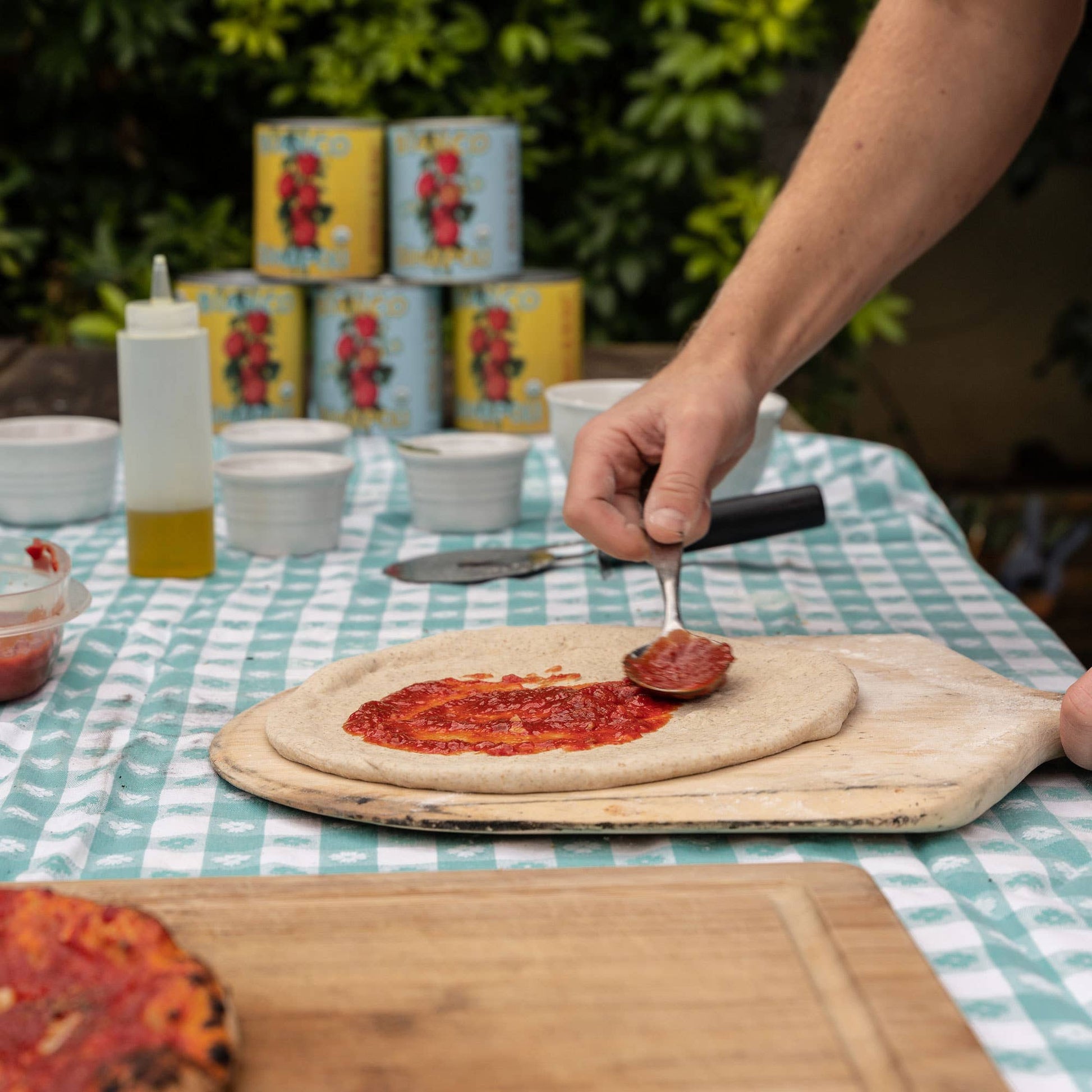 Person spreading pizza sauce on dough with a spoon at a table outdoors.