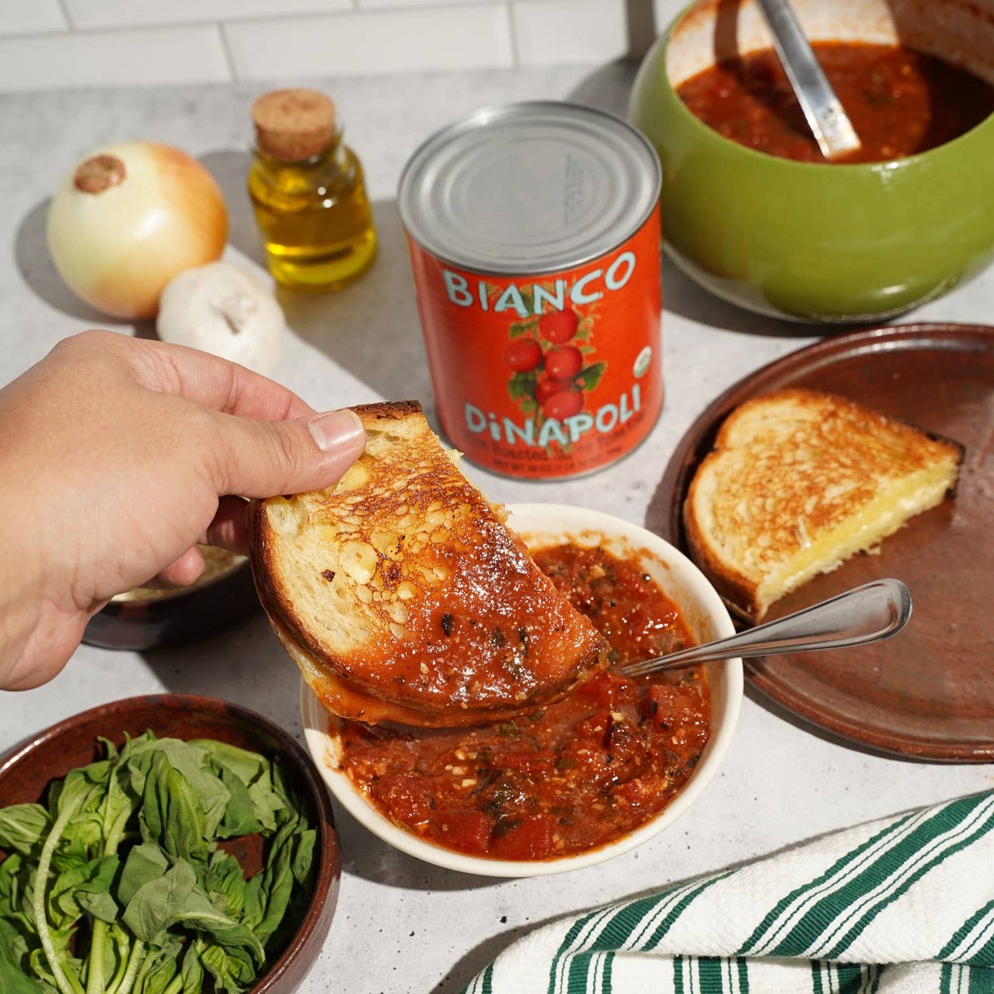 Person spreading Bianco Fire Roasted Tomatoes on toast with various ingredients on a kitchen counter.
