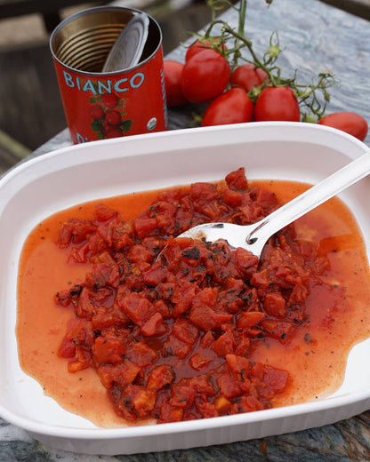A can of Bianco DiNapoli tomatoes with a spoonful of fire roasted chopped tomatoes in a white bowl, surrounded by fresh tomatoes and the can's lid in the background.