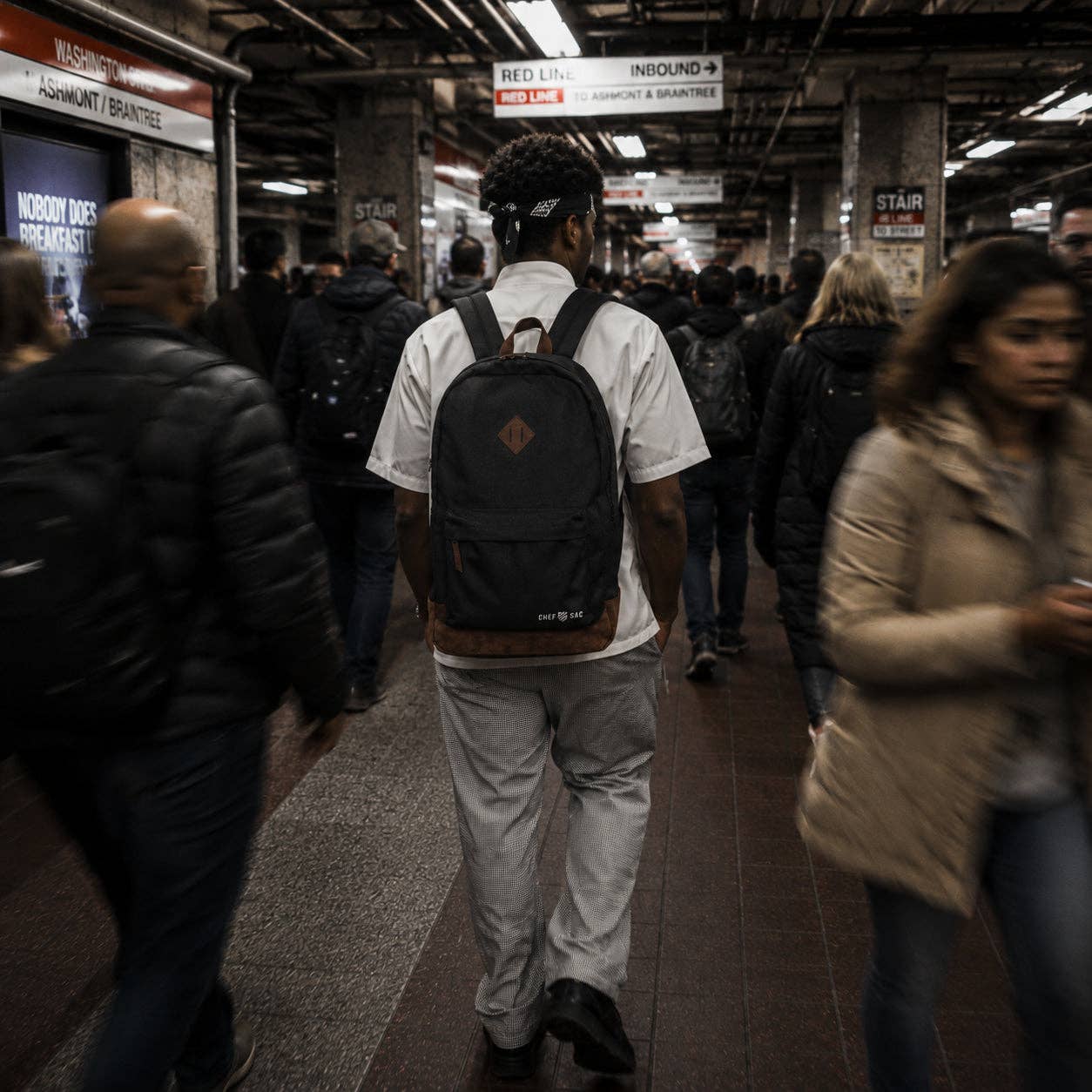 A chef walking away in a crowded subway station, wearing a white t-shirt, light gray pants, and a black backpack with brown trim and a diamond-shaped logo patch.