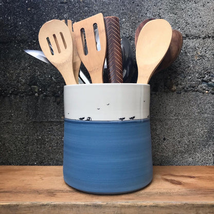 A ceramic utensil holder with a white top section and a blue bottom section, filled with wooden kitchen utensils, set on a wooden surface against a dark textured wall.