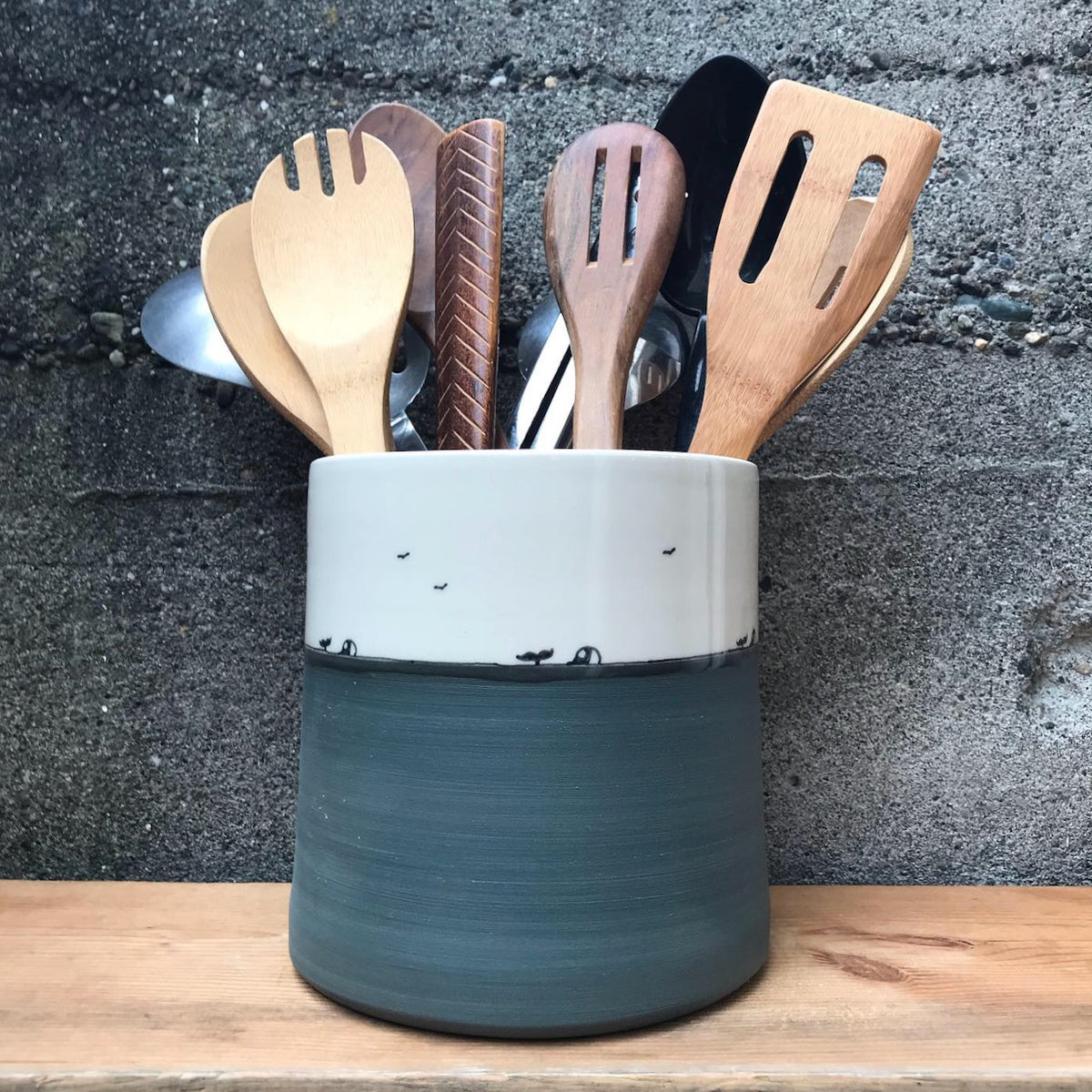 A ceramic kitchen utensil holder with a two-tone design, featuring a white upper section and a dark gray-green lower section, filled with wooden and metal kitchen tools, set on a wooden surface against a dark textured wall.