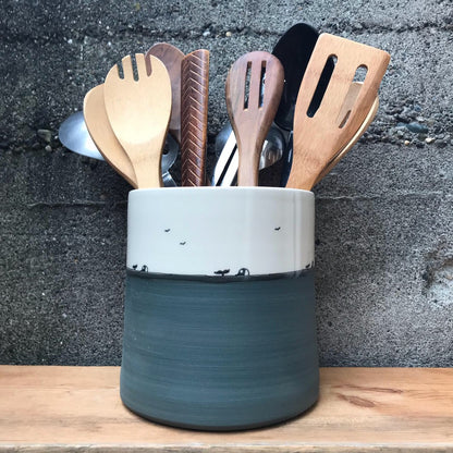 A ceramic kitchen utensil holder with a two-tone design, featuring a white upper section and a dark gray-green lower section, filled with wooden and metal kitchen tools, set on a wooden surface against a dark textured wall.