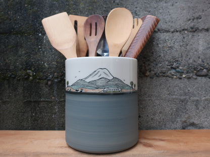 A ceramic kitchen utensil holder with a hand-painted mountain design, filled with wooden spoons and spatulas, set on a wooden surface against a textured gray stone wall.