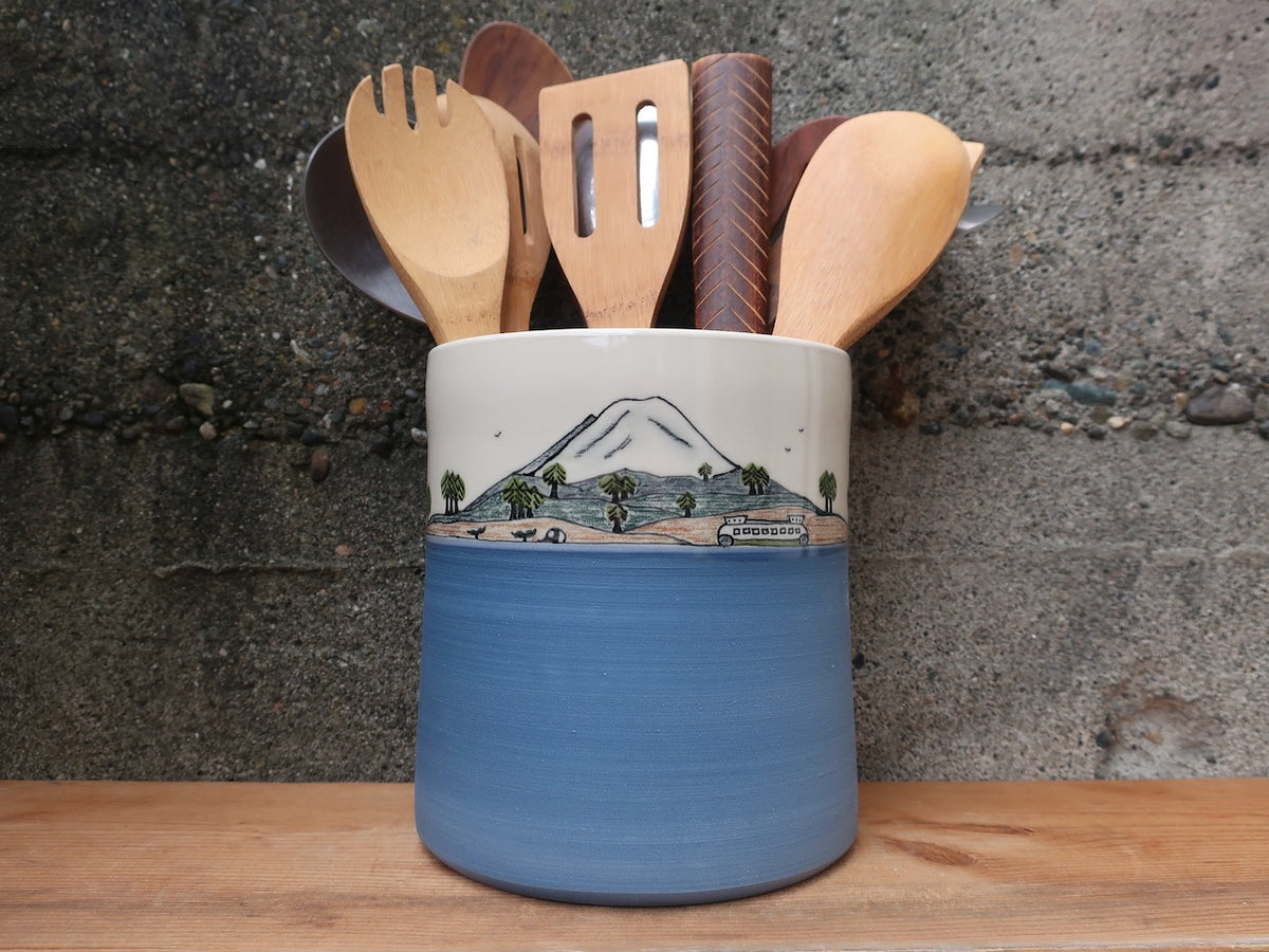 A ceramic kitchen utensil holder with a blue lower half and a hand-painted mountain scene on the upper half, filled with wooden spoons and spatulas, set on a wooden surface against a textured gray wall.