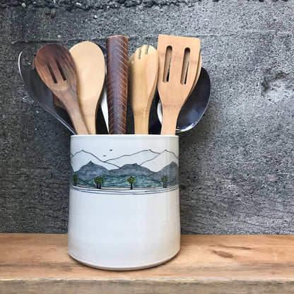 A white ceramic utensil holder with a hand-painted mountain landscape design, filled with wooden and metal kitchen utensils, set on a wooden surface against a textured gray concrete wall.