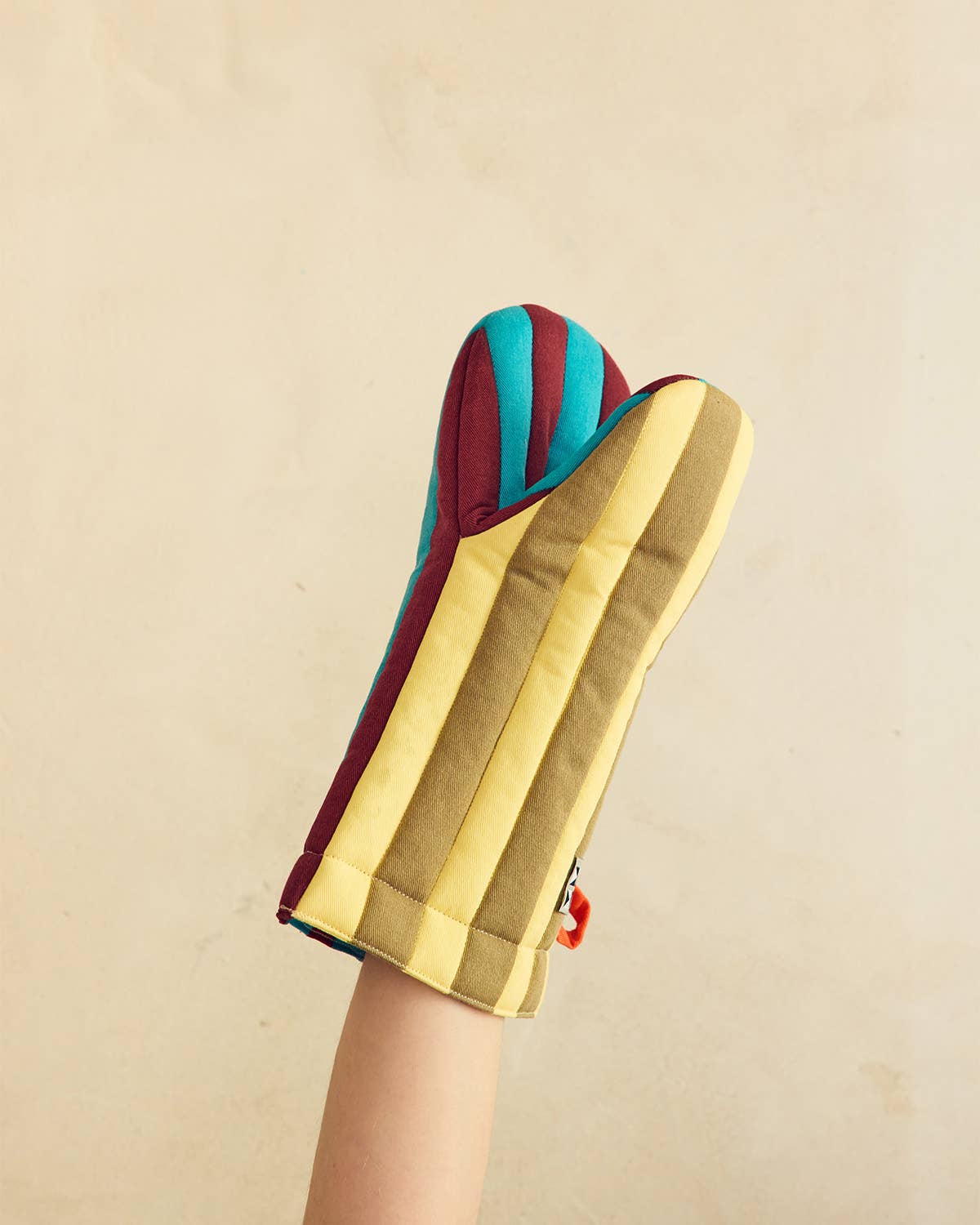 A striped oven mitt held up by a person's arm against a plain beige wall. The mitt features alternating vertical stripes of mustard yellow, olive green, teal blue, and burgundy red.