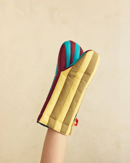 A striped oven mitt held up by a person's arm against a plain beige wall. The mitt features alternating vertical stripes of mustard yellow, olive green, teal blue, and burgundy red.