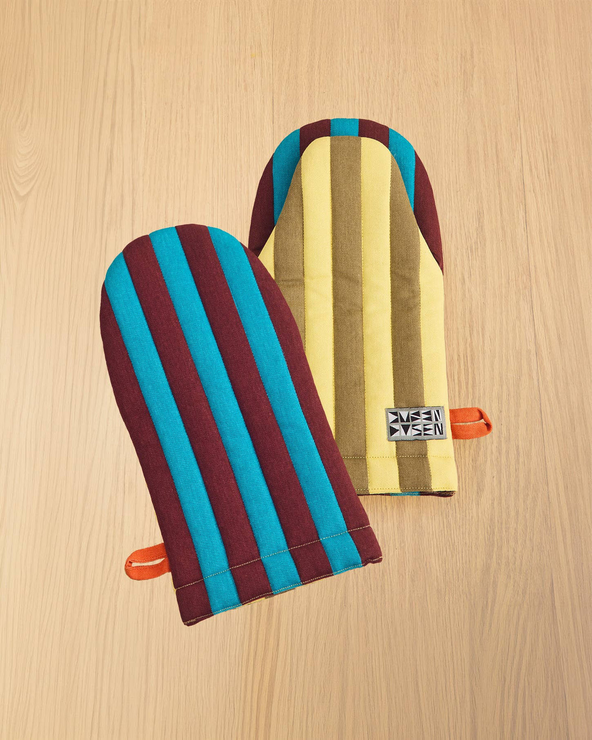 Two striped fabric oven mitts with orange handles, displayed on a light wood grain background. One mitt features vertical stripes of teal blue and dark burgundy, while the other features vertical stripes of yellow, brown, and teal blue.
