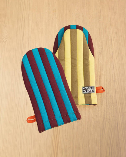 Two striped fabric oven mitts with orange handles, displayed on a light wood grain background. One mitt features vertical stripes of teal blue and dark burgundy, while the other features vertical stripes of yellow, brown, and teal blue.