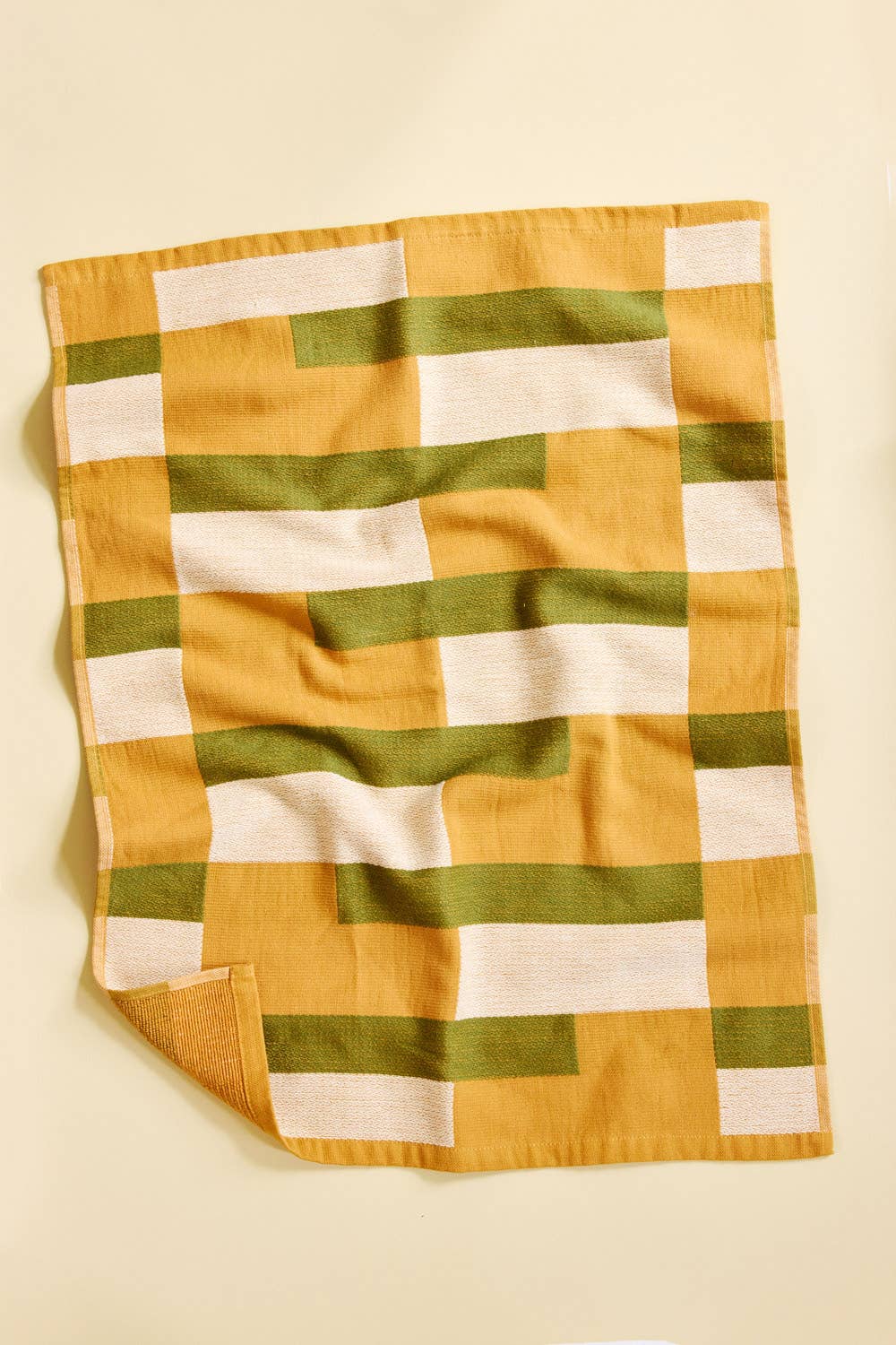 A square cotton kitchen towel with a geometric block pattern in mustard yellow, olive green, and cream, shown flat against a light beige background.