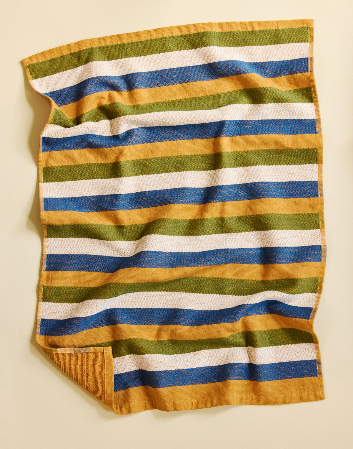 A striped cotton towel with horizontal bands of mustard yellow, olive green, blue, and white, laid flat on a light beige background.