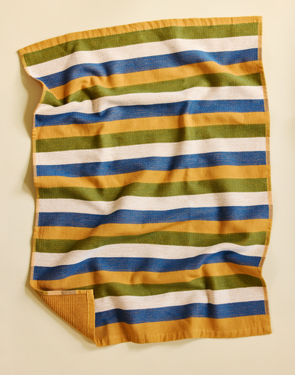 A striped cotton towel with horizontal bands of mustard yellow, olive green, blue, and white, laid flat on a light beige background.