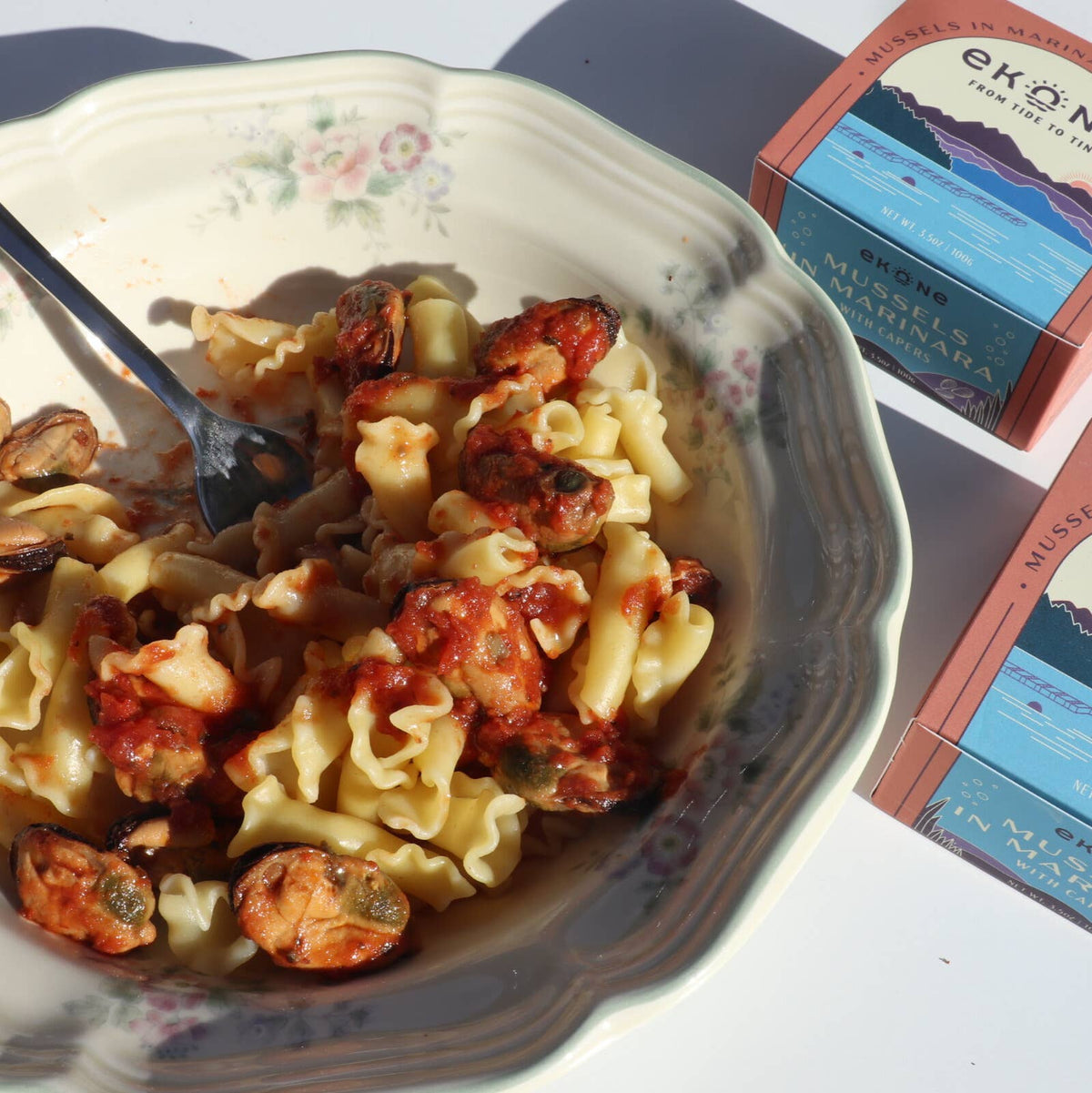 Pasta dish with a fork on a floral-patterned plate, with EKOMAX product packaging in the background.