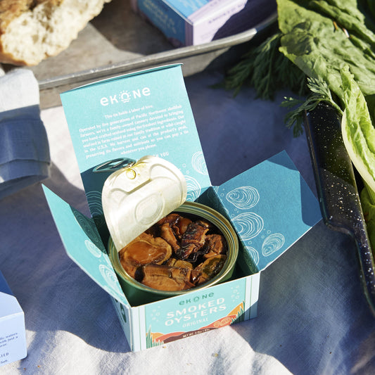 Ekone Smoked Oysters in a teal and white branded box, with the lid propped open to show the product inside and printed packaging details. The scene is styled outdoors on a light fabric surface with fresh herbs and a wooden board in the background.