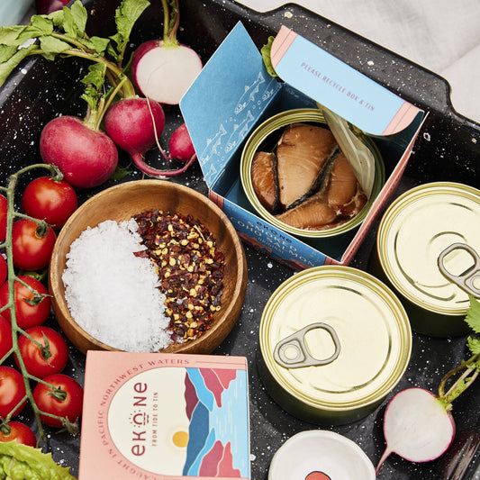 Assorted canned food items, fresh vegetables, and a box of Ekané tea on a black surface.