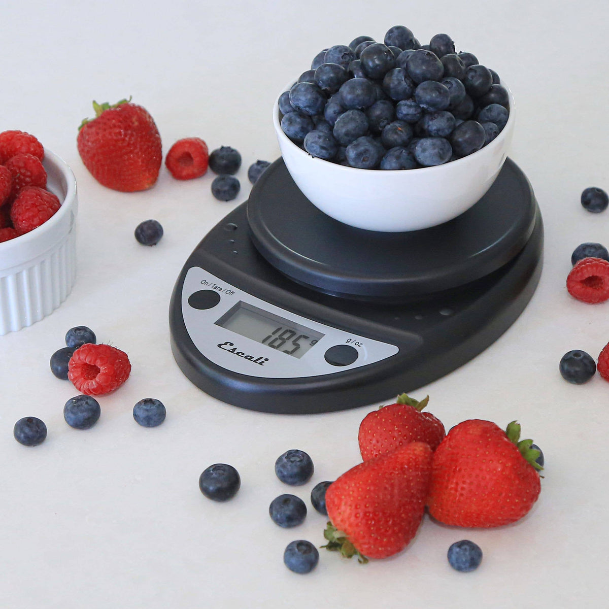 Digital kitchen scale with a bowl of blueberries on a white surface surrounded by strawberries and raspberries.