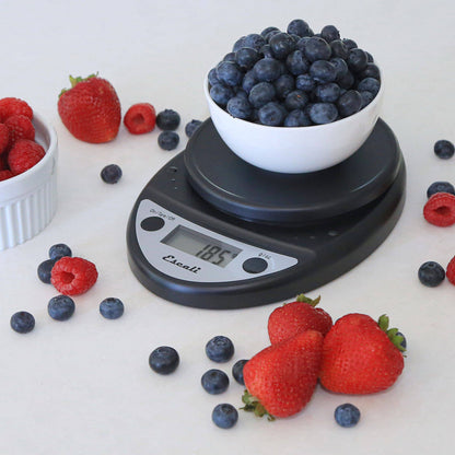 Digital kitchen scale with a bowl of blueberries on a white surface surrounded by strawberries and raspberries.