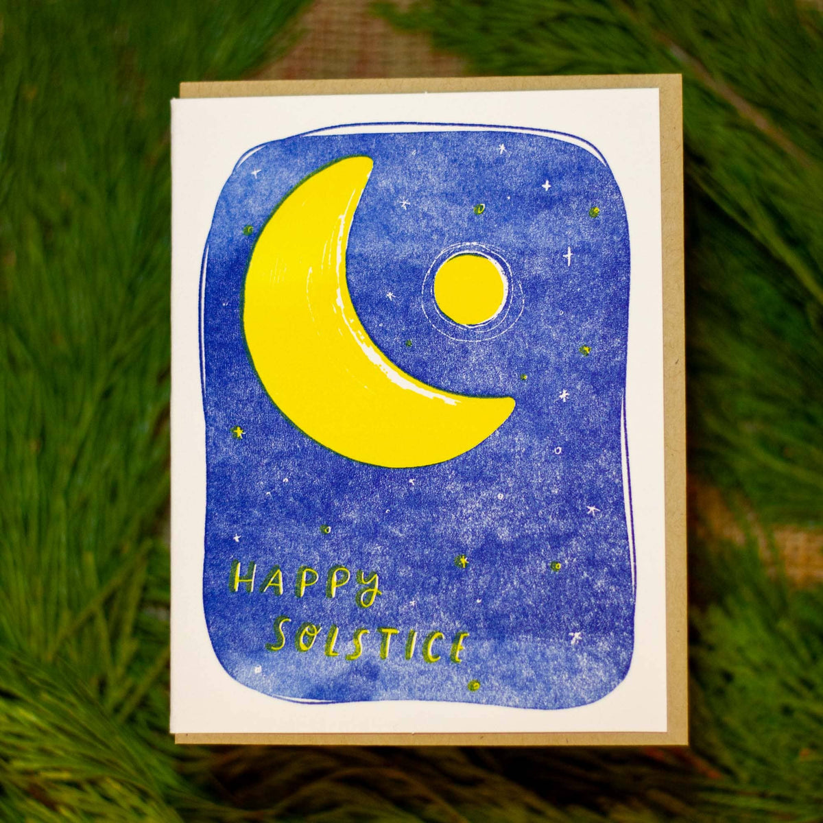 A square greeting card featuring a yellow crescent moon and sun on a blue watercolor night sky with the words 'Happy Solstice', photographed against a background of green pine needles.