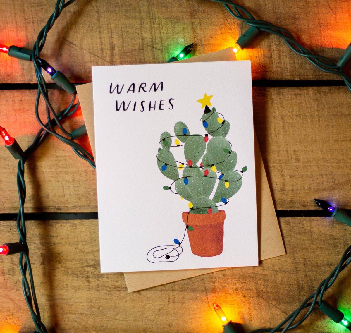Greeting card with a cactus illustration and 'Warm Wishes' text, surrounded by Christmas lights on a wooden surface.