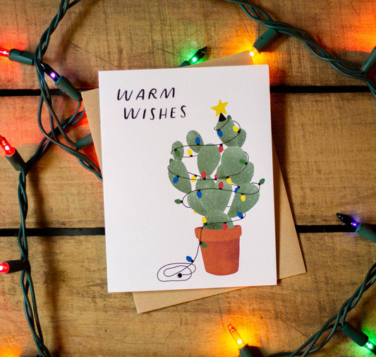 Greeting card with a cactus illustration and 'Warm Wishes' text, surrounded by Christmas lights on a wooden surface.