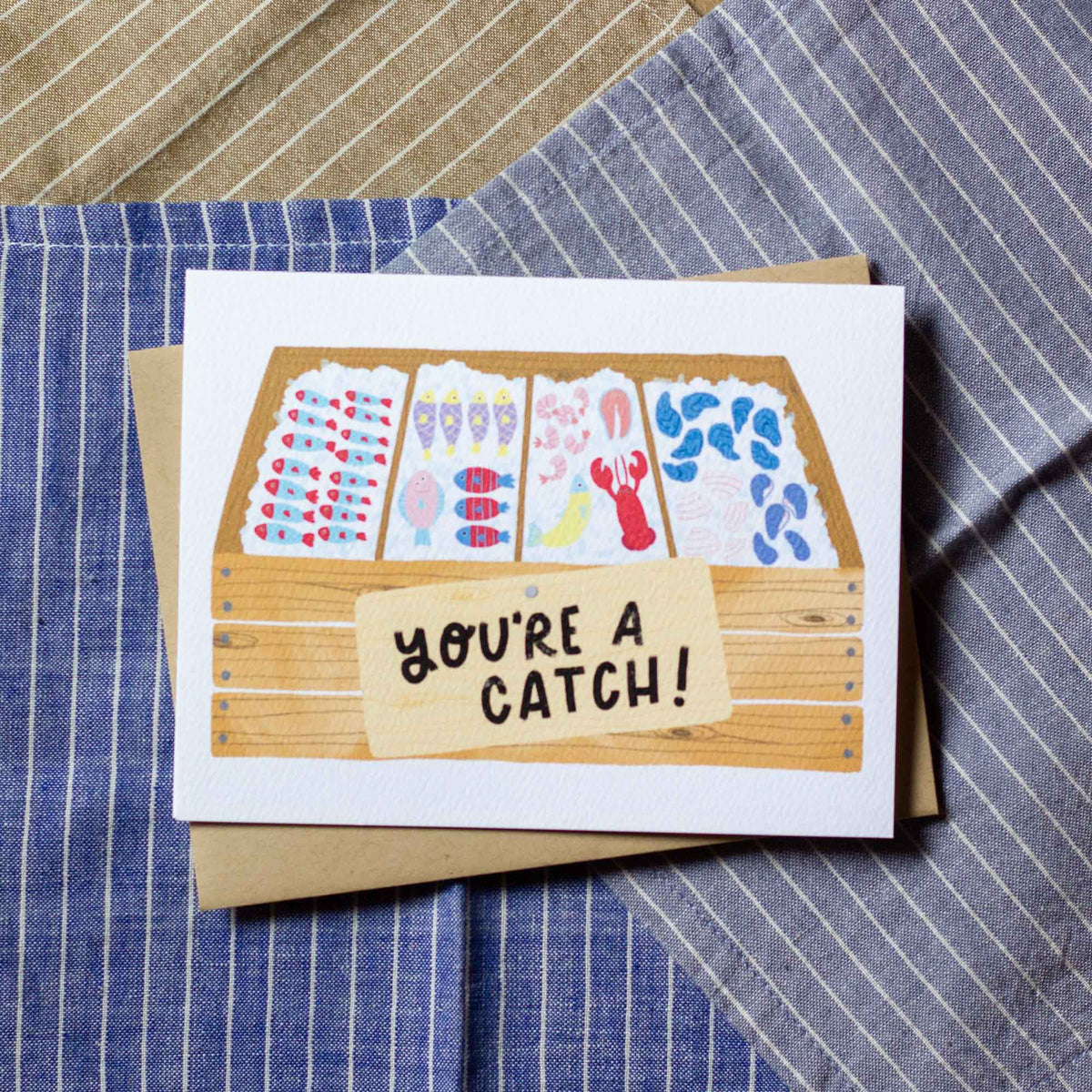 Greeting card with fish market illustration and 'You're a Catch!' text on a striped fabric background