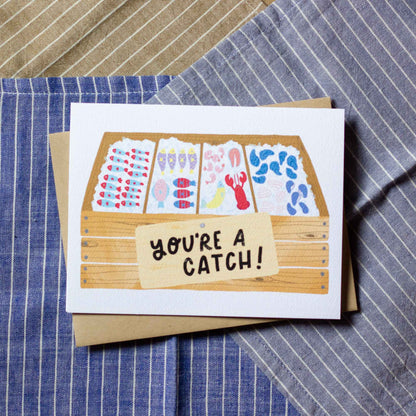 Greeting card with fish market illustration and 'You're a Catch!' text on a striped fabric background