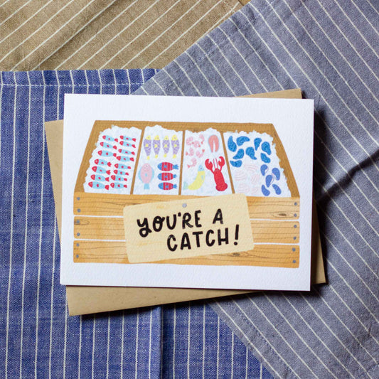 Greeting card with fish market illustration and 'You're a Catch!' text on a striped fabric background