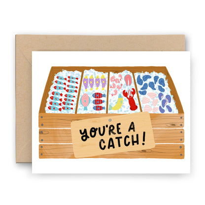 Greeting card with a fish market theme, featuring a sign that reads 'You're a Catch!' and an array of colorful fish-shaped candy items in a wooden crate.