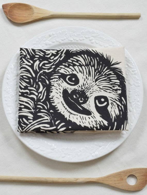A black and white tea towel with a sloth design, displayed on a plate with wooden utensils beside it.