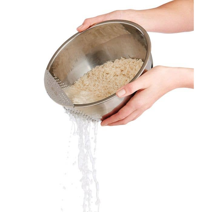 Person rinsing rice in a metal strainer over a bowl on a white background