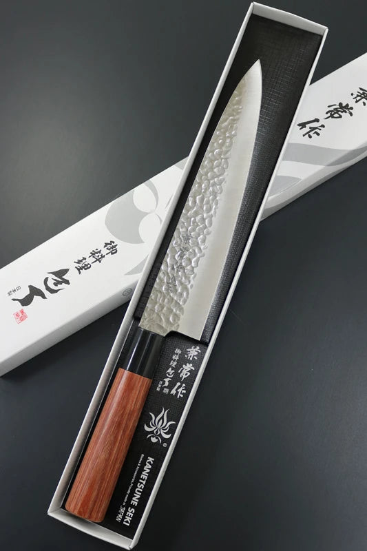 A Japanese kitchen knife with a hammered silver blade and a reddish-brown wooden handle, presented inside an open white and black box with Japanese text on a dark gray background.