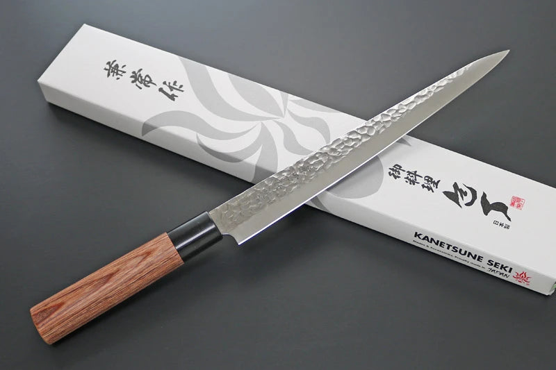 A Japanese kitchen knife with a hammered steel blade and a light brown wooden handle, resting diagonally across its white presentation box with black calligraphy and the brand name Kanetsune Seki.