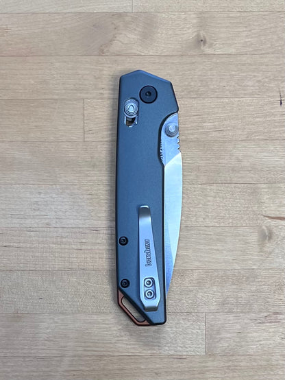 A Kershaw Iridium folding knife with a black anodized aluminum handle and a black-oxide finished blade, viewed from the closed position on a wooden surface.