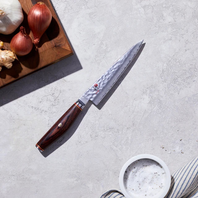 Miyabi Artisan Utility knife on textured white background with cutting board with shallots and garlic.