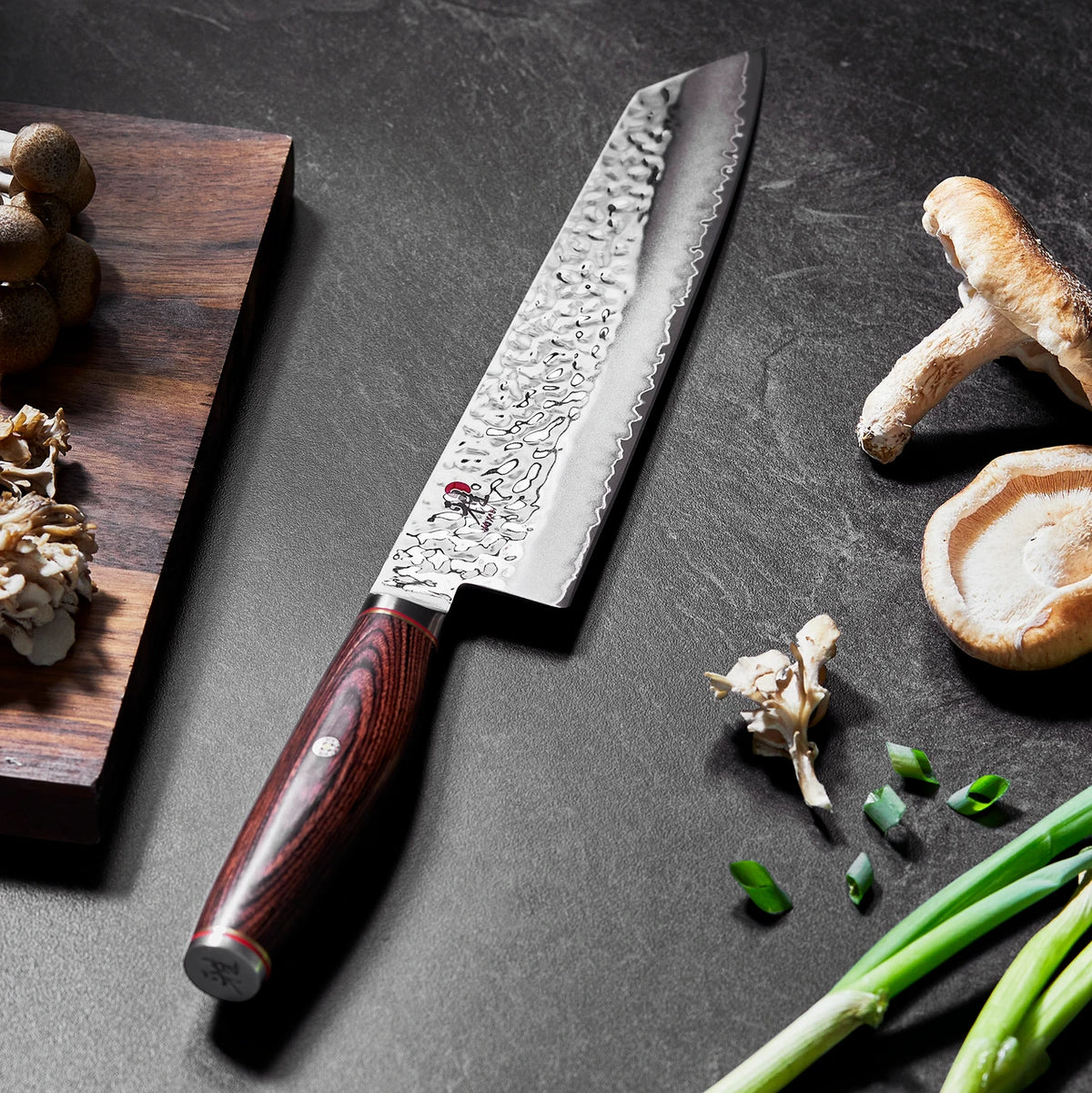 Moody picture of Miyabi Artisan Kiritsuke on  textured black background with mushrooms and scallions.