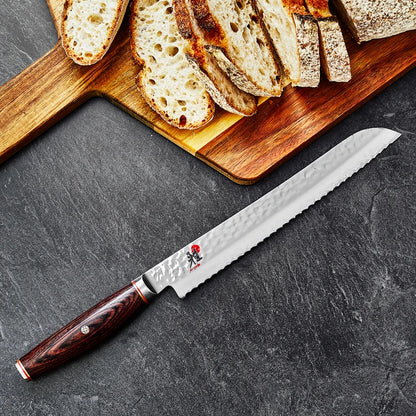 Miyabi Artisan Bread Knife on textured black background with a cutting board with sliced bread.
