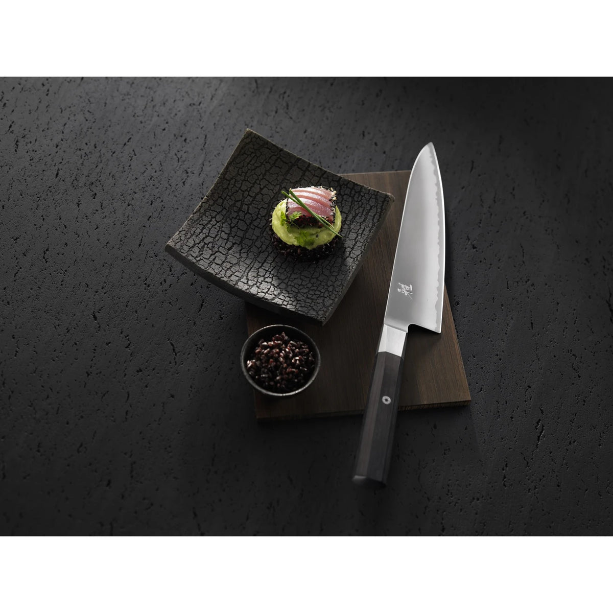 Miyabi chef knife with sushi on textured black background.